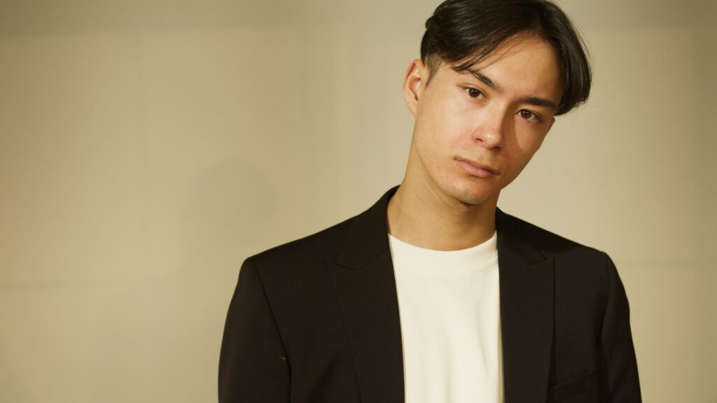 A young man in a black suit jacket, photographed in a studio setting with a neutral expression.