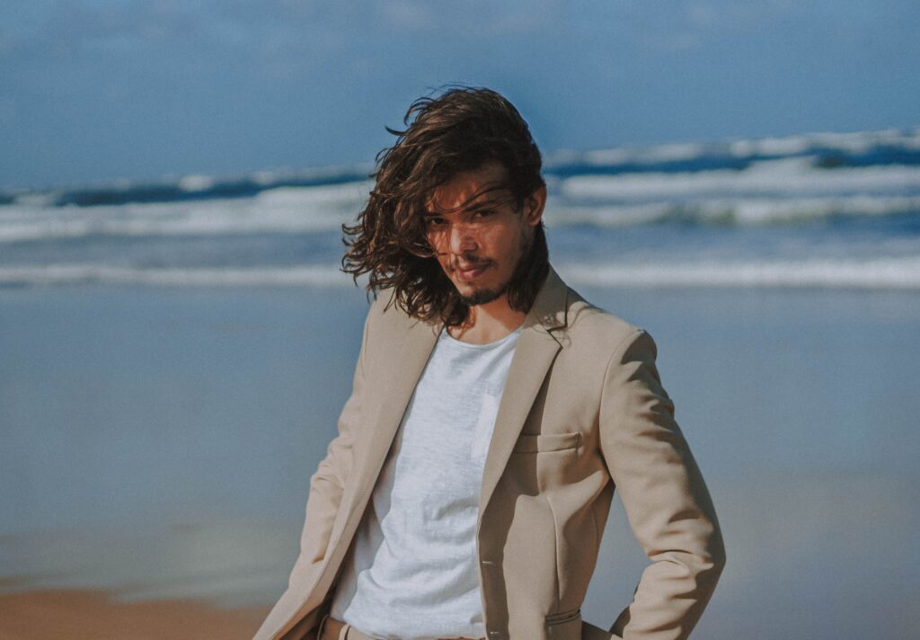 Fashionable man in a beige suit posing on a sunny beach with waves in the background.